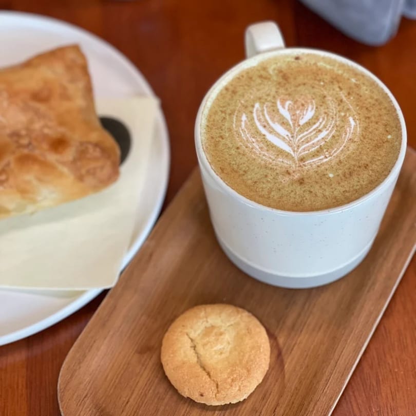 Queen B coffee latte with latte art, a pastry, and a cookie on a wooden tray.