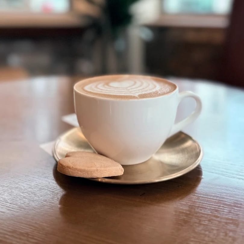 A Queen B latte with heart-shaped art and a cookie on a saucer, on a wooden table.