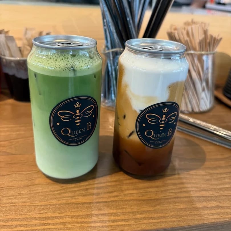 Two Queen B canned drinks, one green matcha and one layered coffee, on a wooden counter.