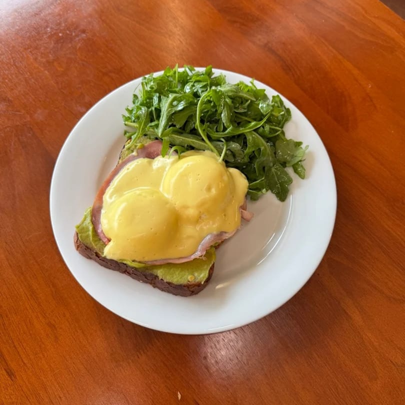 Eggs Benedict with ham and avocado on toast, served with a side of fresh arugula.